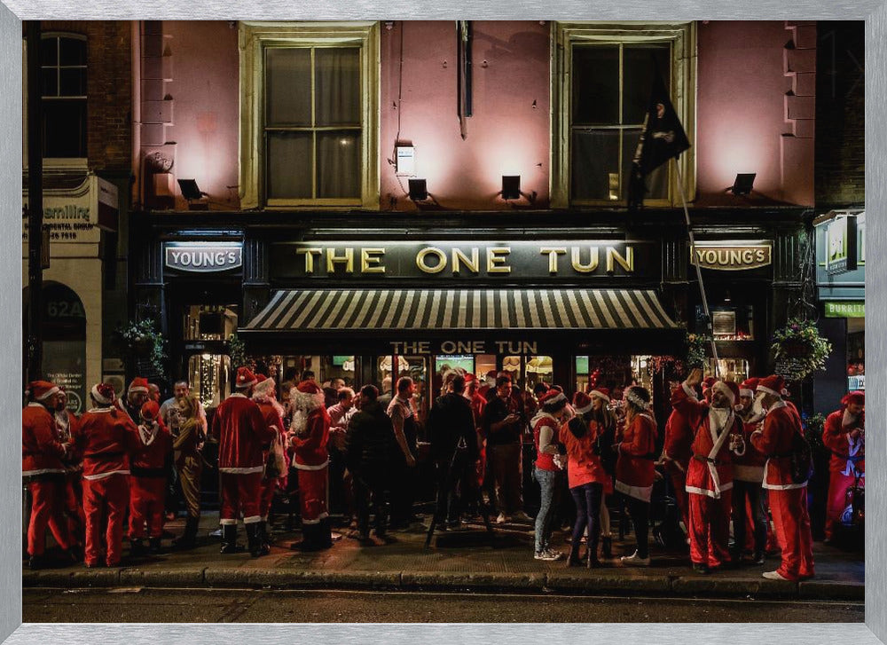A nighttime photograph of a large crowd of people dressed in Santa Claus costumes standing outside a warmly lit pub called 'The One Tun', creating a festive holiday atmosphere on a city street. Print