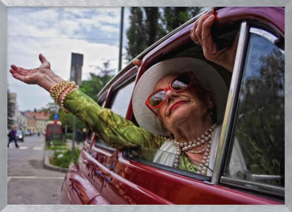 A stylish elderly woman wearing a white hat, red sunglasses, and pearl necklaces leans out of the passenger window of a classic red car. Her arm is outstretched with a joyful expression on her face, enjoying a ride through a city street. Poster