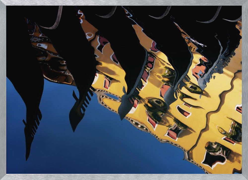 An abstract photograph of a distorted reflection in water, showing a vibrant yellow building against a deep blue sky, with the dark silhouettes of several Venetian gondola prows cutting across the top of the image. The entire scene is enclosed in a silver picture frame. Print
