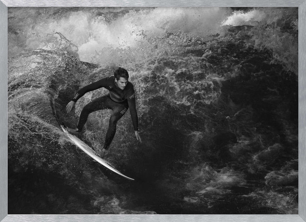 A dramatic black and white photograph of a surfer in a wetsuit skillfully riding down the face of a large, turbulent wave. The image is enclosed in a silver frame. Wall Art