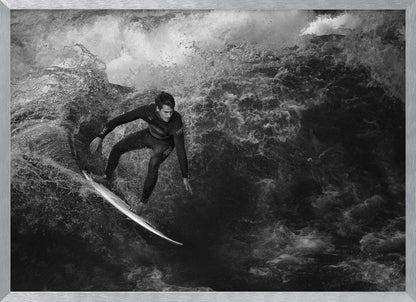 A dramatic black and white photograph of a surfer in a wetsuit skillfully riding down the face of a large, turbulent wave. The image is enclosed in a silver frame. Wall Art