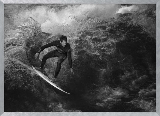 A dramatic black and white photograph of a surfer in a wetsuit skillfully riding down the face of a large, turbulent wave. The image is enclosed in a silver frame. Wall Art