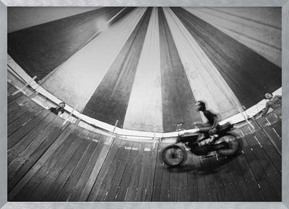 A dynamic, wide-angle black and white photograph of a motorcycle rider performing a stunt in a motordrome, commonly known as the 'wall of death'. The rider is a blur of motion as they speed along the steep, curved wooden walls. The perspective looks up towards the striped canvas top of the tent, with a few spectators visible watching from the railing at the top edge. The entire image is enclosed in a silver frame. Print