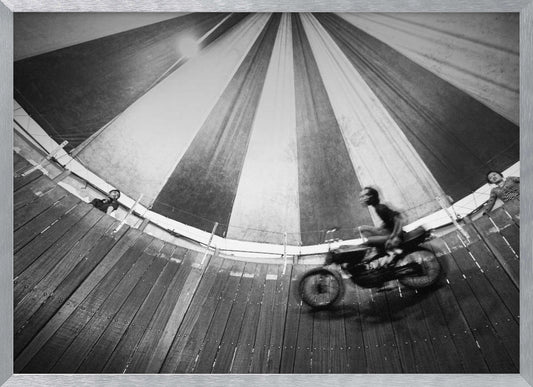A dynamic, wide-angle black and white photograph of a motorcycle rider performing a stunt in a motordrome, commonly known as the 'wall of death'. The rider is a blur of motion as they speed along the steep, curved wooden walls. The perspective looks up towards the striped canvas top of the tent, with a few spectators visible watching from the railing at the top edge. The entire image is enclosed in a silver frame. Print