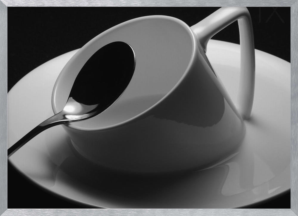 A dramatic black and white close-up photograph of a white porcelain coffee cup tilted on its saucer, with a reflective spoon resting inside. The image has high contrast against a dark background and is presented within a silver-colored frame. Decor