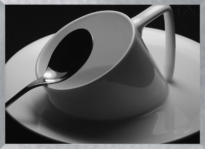 A dramatic black and white close-up photograph of a white porcelain coffee cup tilted on its saucer, with a reflective spoon resting inside. The image has high contrast against a dark background and is presented within a silver-colored frame. Decor