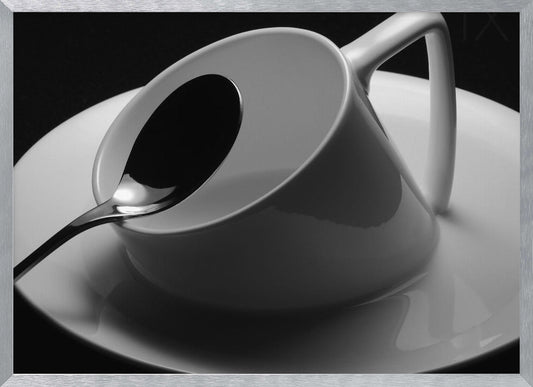 A dramatic black and white close-up photograph of a white porcelain coffee cup tilted on its saucer, with a reflective spoon resting inside. The image has high contrast against a dark background and is presented within a silver-colored frame. Decor