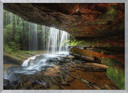A long-exposure photograph taken from underneath a rock overhang, looking out through a cascading waterfall. The water appears silky as it flows over mossy rocks in a stream below. The scene is framed by a silver border. Print