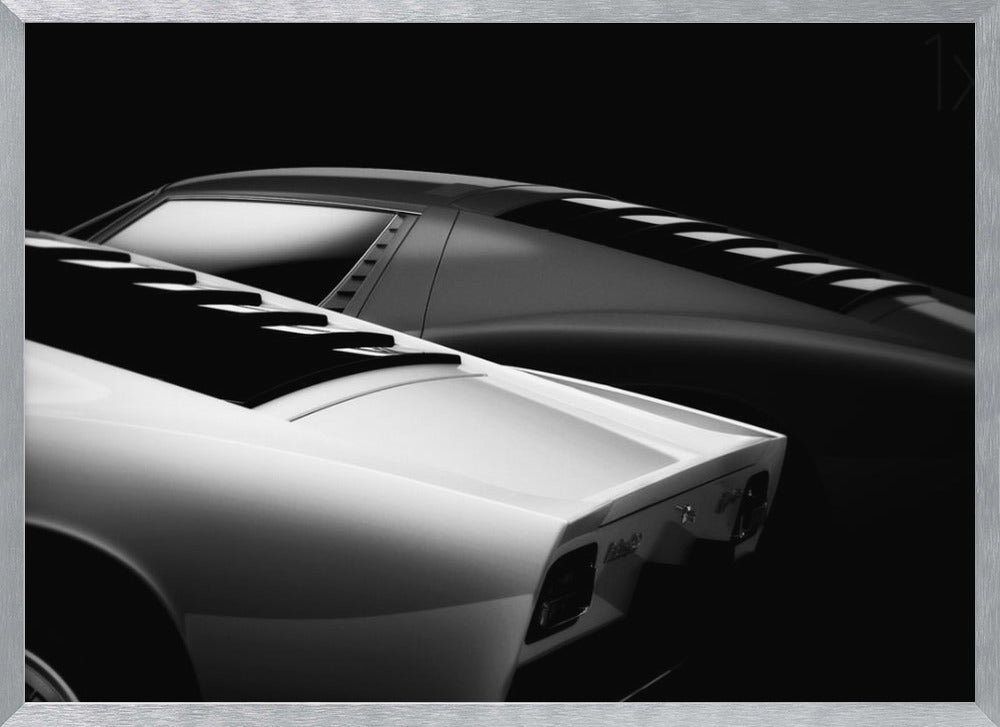 A dramatic black and white studio photograph showcasing the sleek, curved lines of two classic sports cars, with a white car in the foreground and a dark one in the background, set against a black backdrop. Wall Art