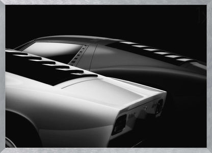 A dramatic black and white studio photograph showcasing the sleek, curved lines of two classic sports cars, with a white car in the foreground and a dark one in the background, set against a black backdrop. Wall Art