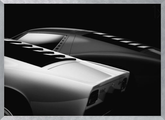 A dramatic black and white studio photograph showcasing the sleek, curved lines of two classic sports cars, with a white car in the foreground and a dark one in the background, set against a black backdrop. Wall Art