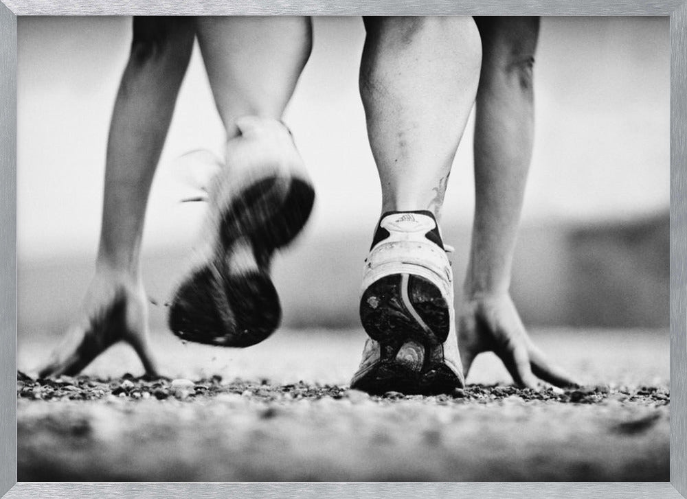 A low-angle, black and white action shot of a runner's legs and hands at the starting position on a gravel track. One foot is planted firmly while the other is in motion, blurred as they push off to start a race. Decor