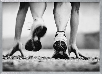 A low-angle, black and white action shot of a runner's legs and hands at the starting position on a gravel track. One foot is planted firmly while the other is in motion, blurred as they push off to start a race. Decor