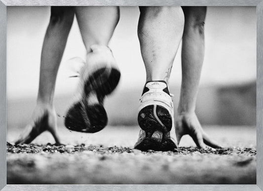 A low-angle, black and white action shot of a runner's legs and hands at the starting position on a gravel track. One foot is planted firmly while the other is in motion, blurred as they push off to start a race. Decor