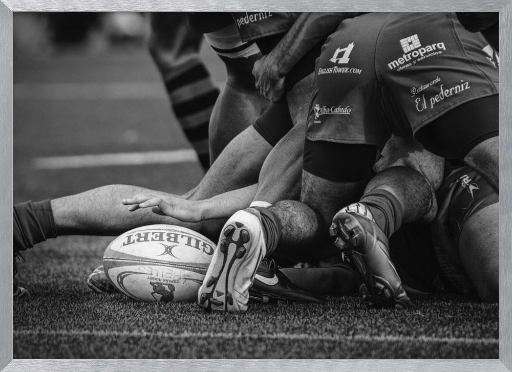A dynamic black and white, low-angle photograph capturing the intensity of a rugby ruck. A Gilbert rugby ball sits on the turf in the foreground, surrounded by a tangle of players' muscular legs, arms, and cleats, conveying the raw power and action of the sport. Wall Art