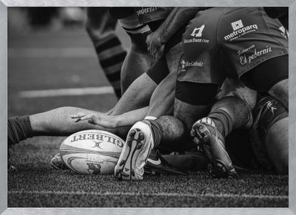 A dynamic black and white, low-angle photograph capturing the intensity of a rugby ruck. A Gilbert rugby ball sits on the turf in the foreground, surrounded by a tangle of players' muscular legs, arms, and cleats, conveying the raw power and action of the sport. Wall Art