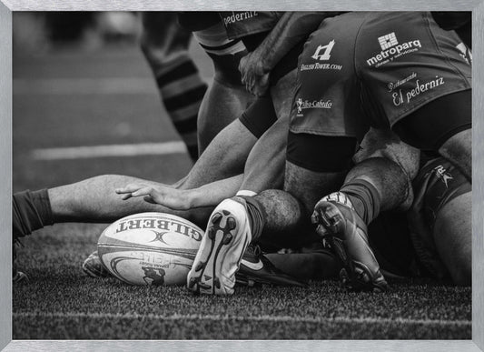 A dynamic black and white, low-angle photograph capturing the intensity of a rugby ruck. A Gilbert rugby ball sits on the turf in the foreground, surrounded by a tangle of players' muscular legs, arms, and cleats, conveying the raw power and action of the sport. Wall Art