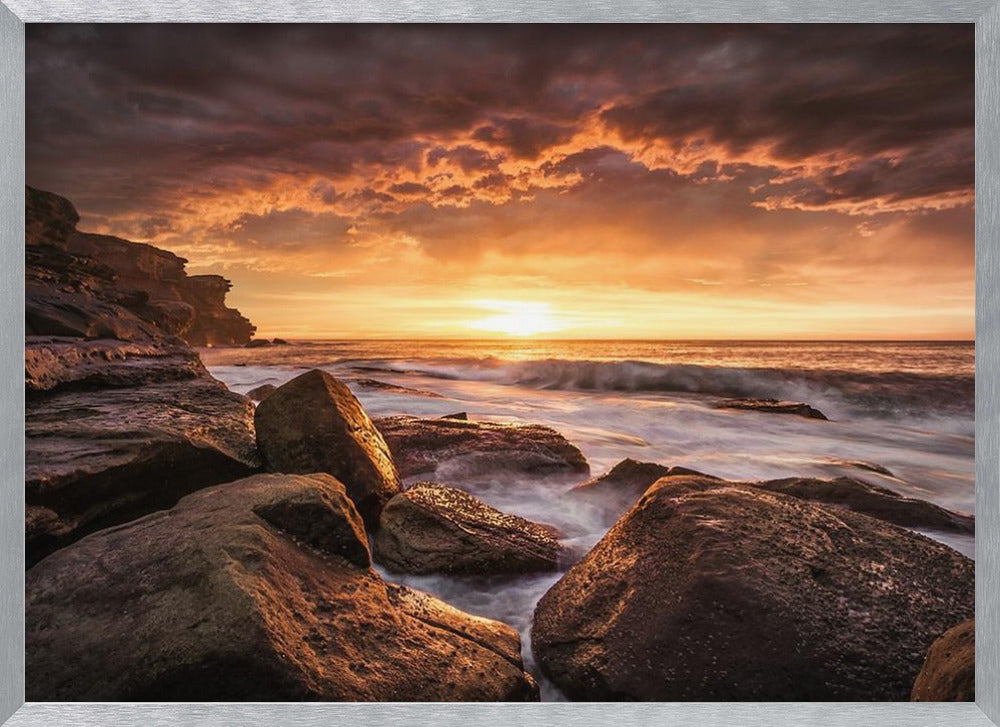 A framed photograph of a dramatic ocean sunset, with fiery orange clouds filling the sky. Large, dark rocks sit on the shore as long-exposure waves create a misty effect around them. Print
