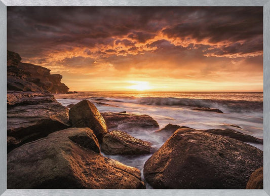 A framed photograph of a dramatic ocean sunset, with fiery orange clouds filling the sky. Large, dark rocks sit on the shore as long-exposure waves create a misty effect around them. Print