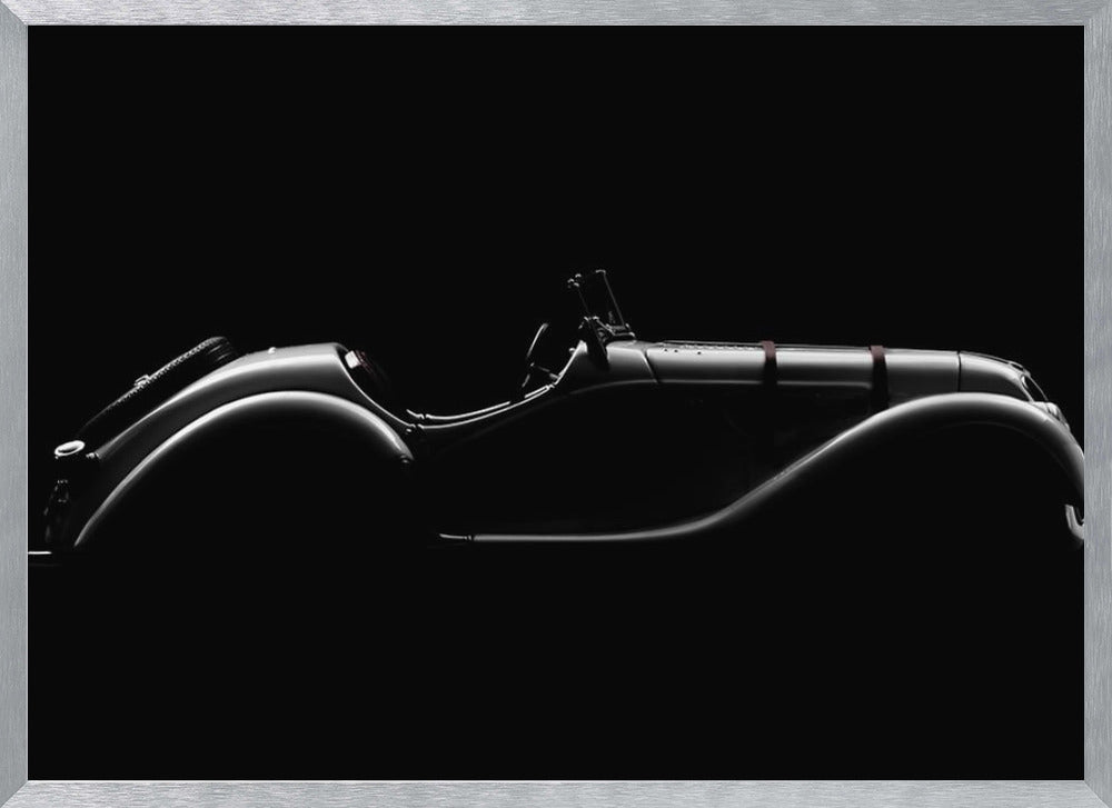 A dramatic black and white photograph showcasing the elegant silhouette of a vintage roadster. The side profile of the classic car is highlighted by subtle light, emphasizing its long hood and sweeping fenders against a solid black background, all within a brushed silver frame. Print