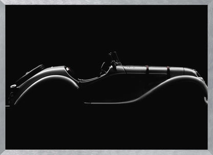 A dramatic black and white photograph showcasing the elegant silhouette of a vintage roadster. The side profile of the classic car is highlighted by subtle light, emphasizing its long hood and sweeping fenders against a solid black background, all within a brushed silver frame. Print