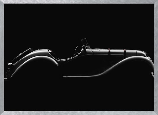 A dramatic black and white photograph showcasing the elegant silhouette of a vintage roadster. The side profile of the classic car is highlighted by subtle light, emphasizing its long hood and sweeping fenders against a solid black background, all within a brushed silver frame. Print