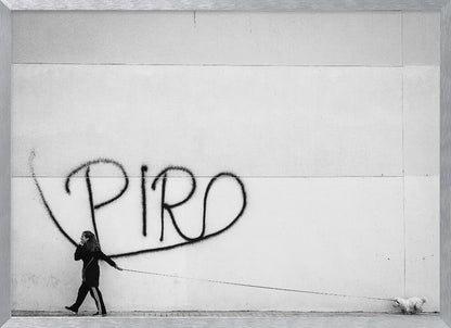 A minimalist black and white photograph of a woman dressed in black walking a small, fluffy white dog on a long leash. They are walking along a sidewalk in front of a large, pale wall with the word 'PIRO' written in black spray paint graffiti. The entire image is enclosed in a light grey frame. Poster