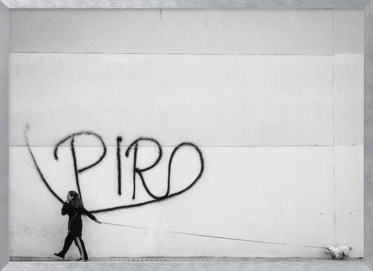 A minimalist black and white photograph of a woman dressed in black walking a small, fluffy white dog on a long leash. They are walking along a sidewalk in front of a large, pale wall with the word 'PIRO' written in black spray paint graffiti. The entire image is enclosed in a light grey frame. Poster