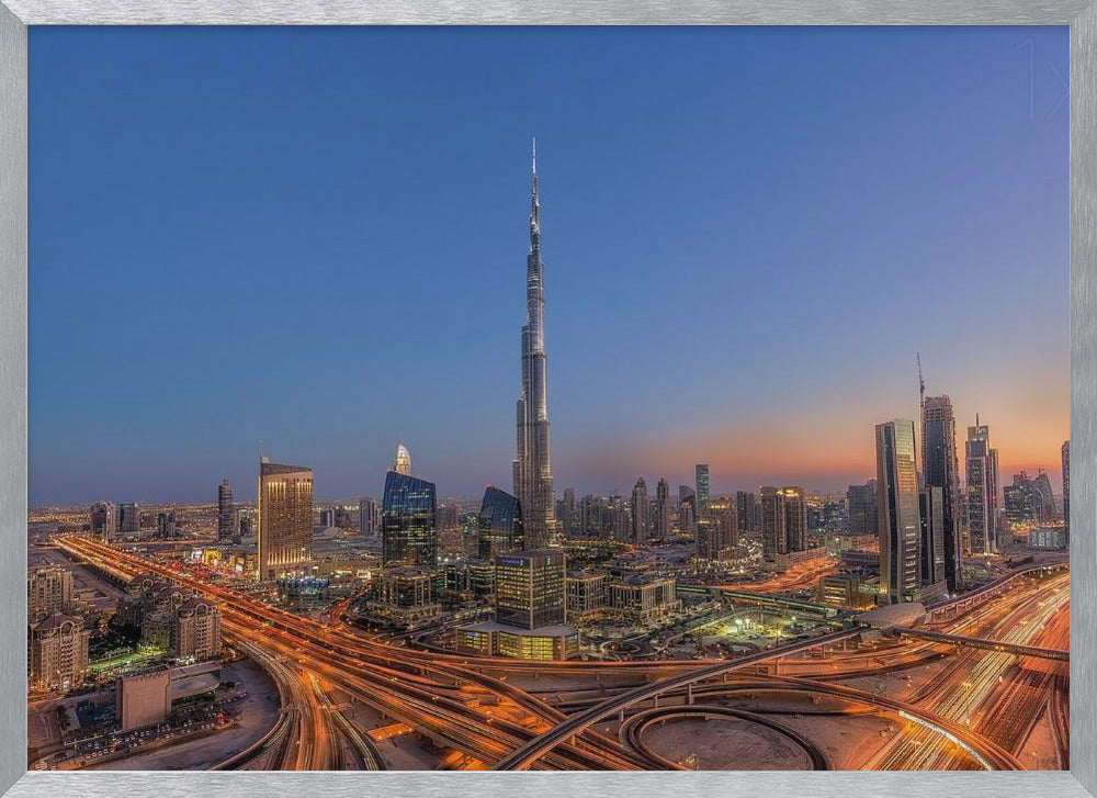 An aerial photograph of the Dubai skyline at dusk, centered on the illuminated Burj Khalifa. The sky transitions from deep blue to warm orange at the horizon, while a complex network of highways below glows with the light trails of traffic. Artwork