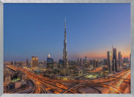 An aerial photograph of the Dubai skyline at dusk, centered on the illuminated Burj Khalifa. The sky transitions from deep blue to warm orange at the horizon, while a complex network of highways below glows with the light trails of traffic. Artwork