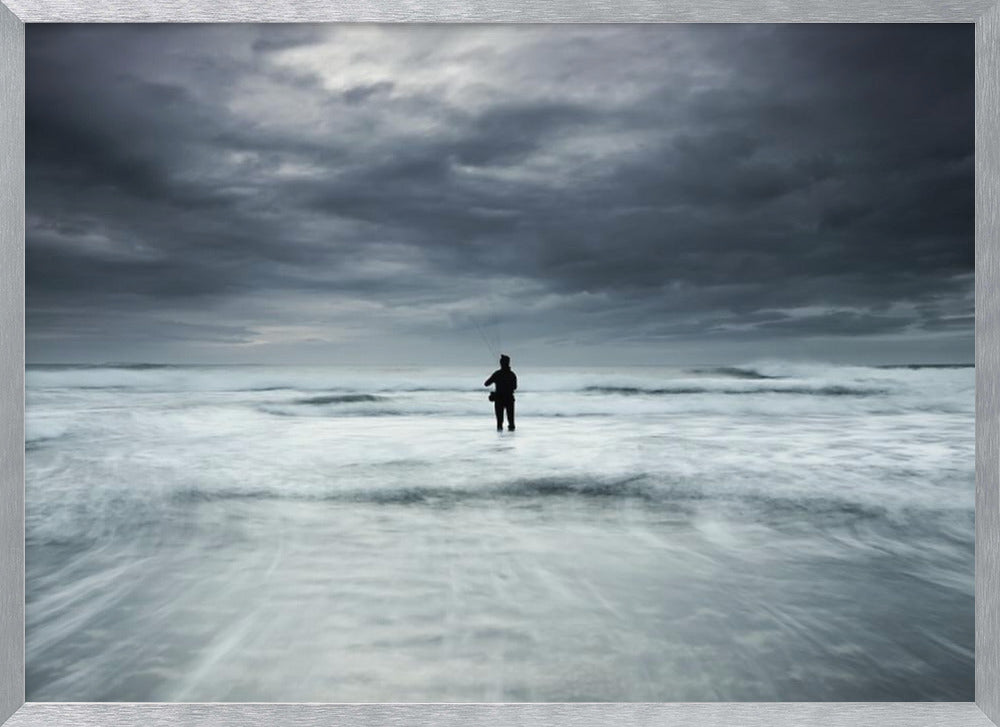 A moody, long-exposure photograph of a silhouetted fisherman standing in the churning white surf of the ocean under a dark, stormy sky, presented in a silver frame. Wall Art