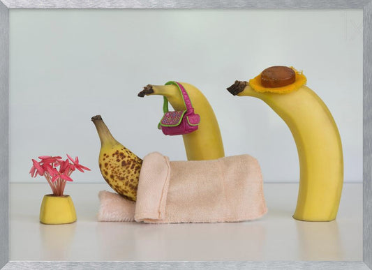 A whimsical still life photo featuring three bananas personified as a family or friends. One overripe, spotted banana lies down, covered by a pink towel, while another banana with a tiny pink purse and a third with a small hat seem to be visiting. A small vase with pink flowers sits nearby on the white surface. Poster
