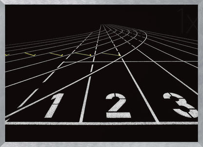 A framed, high-contrast graphic of a black running track from a low-angle perspective at the starting line. White lines delineate the lanes, with the numbers 1, 2, and 3 stenciled in the foreground. The track curves and recedes into the distance, creating a strong sense of perspective. Decor