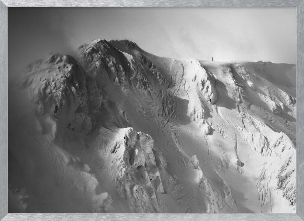 A dramatic black and white photograph of a person standing alone on the ridge of a massive, snow-covered mountain, emphasizing the scale and solitude of the vast, rugged landscape, all presented in a silver frame. Wall Art