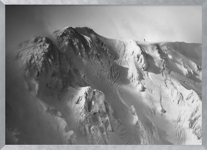 A dramatic black and white photograph of a person standing alone on the ridge of a massive, snow-covered mountain, emphasizing the scale and solitude of the vast, rugged landscape, all presented in a silver frame. Wall Art