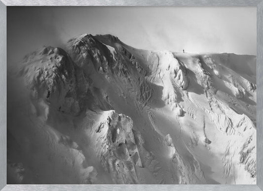A dramatic black and white photograph of a person standing alone on the ridge of a massive, snow-covered mountain, emphasizing the scale and solitude of the vast, rugged landscape, all presented in a silver frame. Wall Art