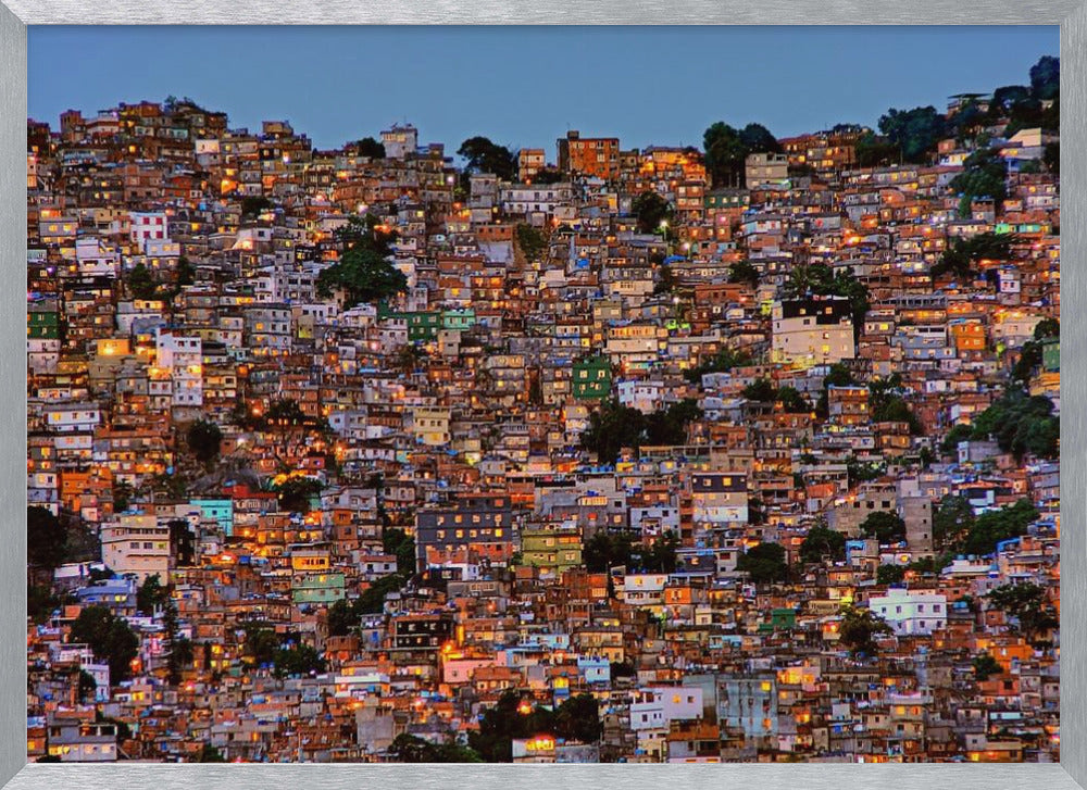 A photograph of a densely packed favela built on a hillside at dusk. The numerous small, colorful houses are illuminated from within, casting a warm orange glow that contrasts with the deep blue of the twilight sky. The image is presented within a silver picture frame. Artwork
