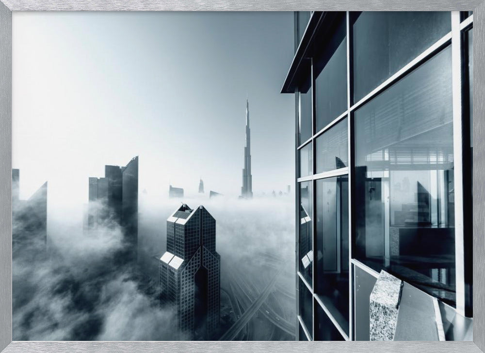A framed black and white photograph with a cool blue tint, depicting the Dubai skyline from a high-rise window, with skyscrapers including the Burj Khalifa piercing through a thick blanket of fog. Artwork