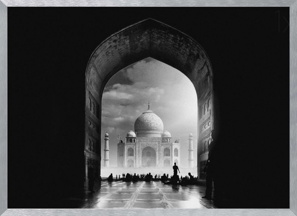 A dramatic black and white photograph of the Taj Mahal seen through a large, dark archway which frames the view. The iconic mausoleum is in the distance under a cloudy sky, and the silhouettes of many people are scattered on the wet, reflective ground in the foreground. Decor