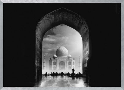 A dramatic black and white photograph of the Taj Mahal seen through a large, dark archway which frames the view. The iconic mausoleum is in the distance under a cloudy sky, and the silhouettes of many people are scattered on the wet, reflective ground in the foreground. Decor