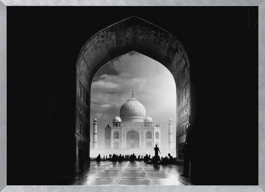 A dramatic black and white photograph of the Taj Mahal seen through a large, dark archway which frames the view. The iconic mausoleum is in the distance under a cloudy sky, and the silhouettes of many people are scattered on the wet, reflective ground in the foreground. Decor