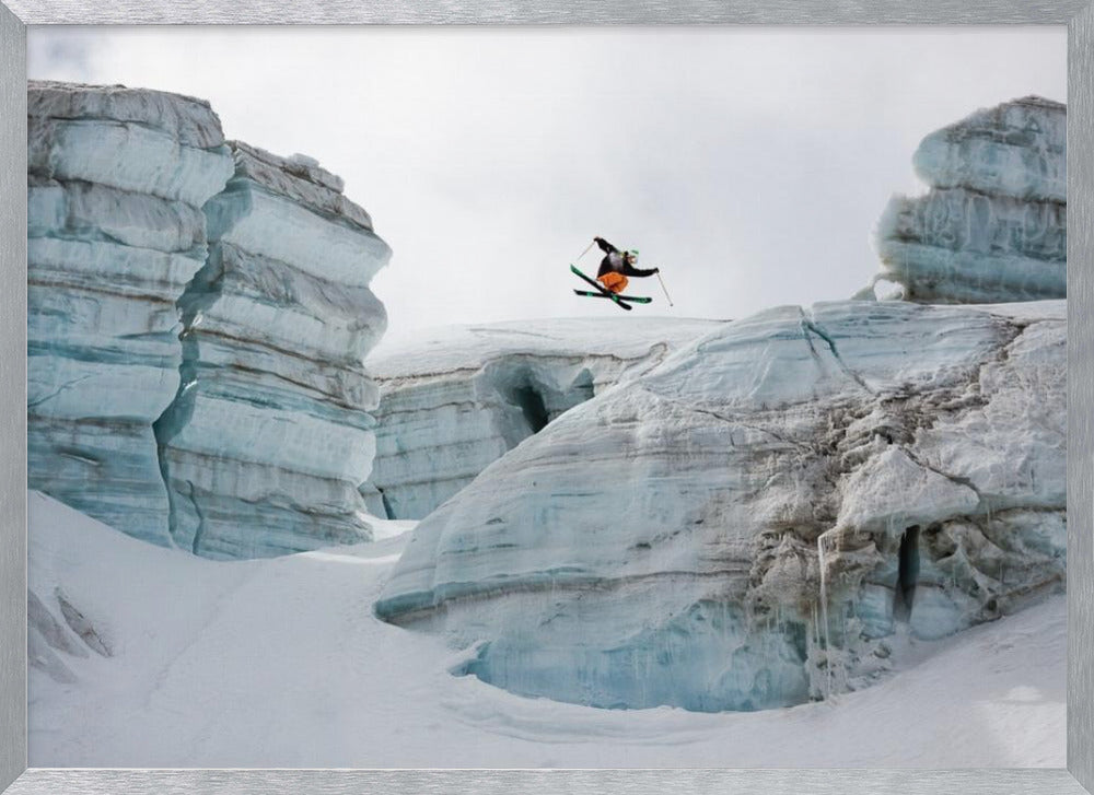 A framed photo of a skier in mid-air, jumping over a snowy crevasse between large, layered glacier formations under an overcast sky. The skier wears a black top and orange pants. Wall Art