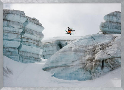 A framed photo of a skier in mid-air, jumping over a snowy crevasse between large, layered glacier formations under an overcast sky. The skier wears a black top and orange pants. Wall Art