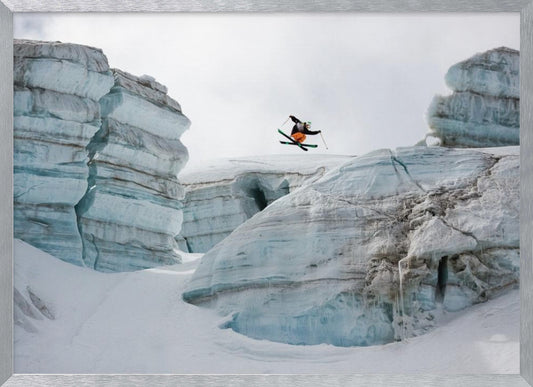 A framed photo of a skier in mid-air, jumping over a snowy crevasse between large, layered glacier formations under an overcast sky. The skier wears a black top and orange pants. Wall Art