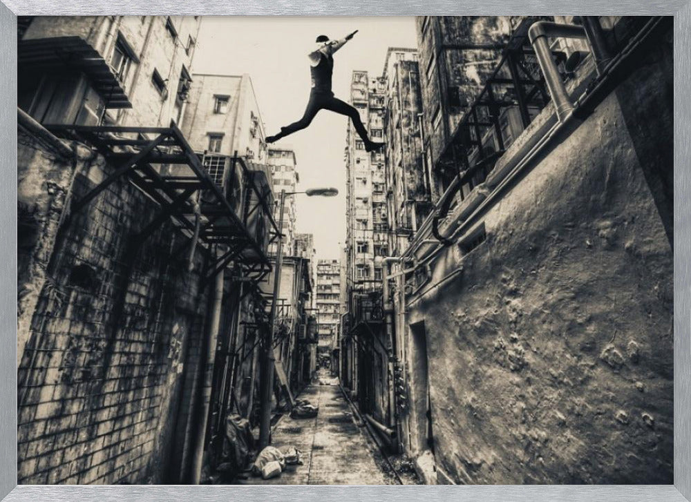 A dramatic black and white photograph of a man in mid-air, leaping athletically across a narrow, gritty urban alleyway. The low-angle shot looks up between tall, weathered buildings, emphasizing the height of the jump. The image is presented in a silver frame. Wall Art