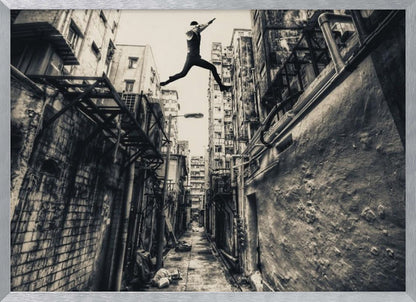 A dramatic black and white photograph of a man in mid-air, leaping athletically across a narrow, gritty urban alleyway. The low-angle shot looks up between tall, weathered buildings, emphasizing the height of the jump. The image is presented in a silver frame. Wall Art