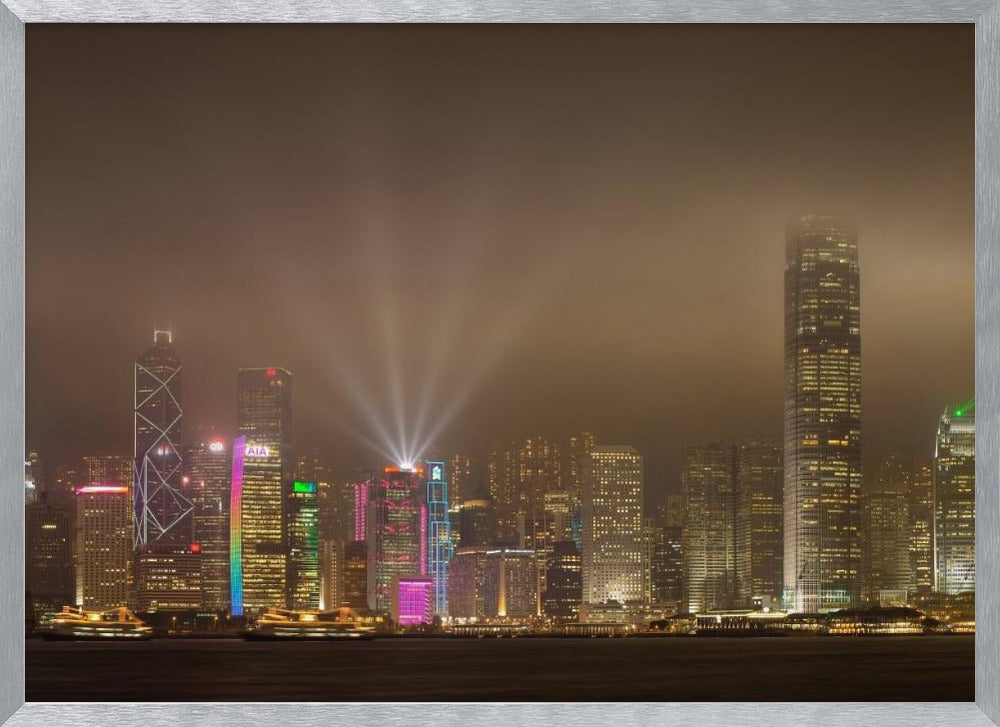 A framed photograph of the Hong Kong skyline at night, viewed from across the water. The skyscrapers are brilliantly lit with yellow, white, and colorful lights, and beams of light shoot up into the foggy, glowing sky. Decor