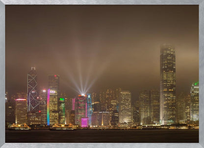 A framed photograph of the Hong Kong skyline at night, viewed from across the water. The skyscrapers are brilliantly lit with yellow, white, and colorful lights, and beams of light shoot up into the foggy, glowing sky. Decor