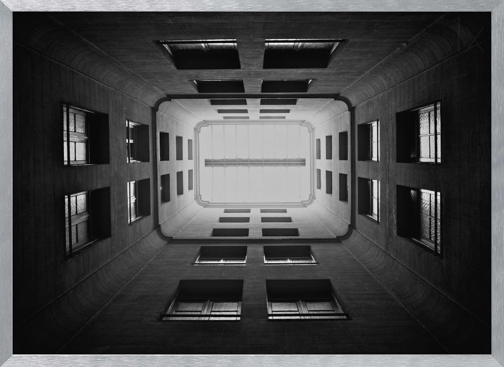 A symmetrical, black and white photograph looking directly up from the base of a building's interior courtyard. The dark walls rise up, lined with rows of windows, creating a powerful sense of perspective that draws the eye to the bright rectangle of the sky or a skylight at the top. The image is enclosed in a simple silver frame. Poster