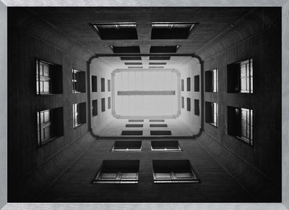 A symmetrical, black and white photograph looking directly up from the base of a building's interior courtyard. The dark walls rise up, lined with rows of windows, creating a powerful sense of perspective that draws the eye to the bright rectangle of the sky or a skylight at the top. The image is enclosed in a simple silver frame. Poster